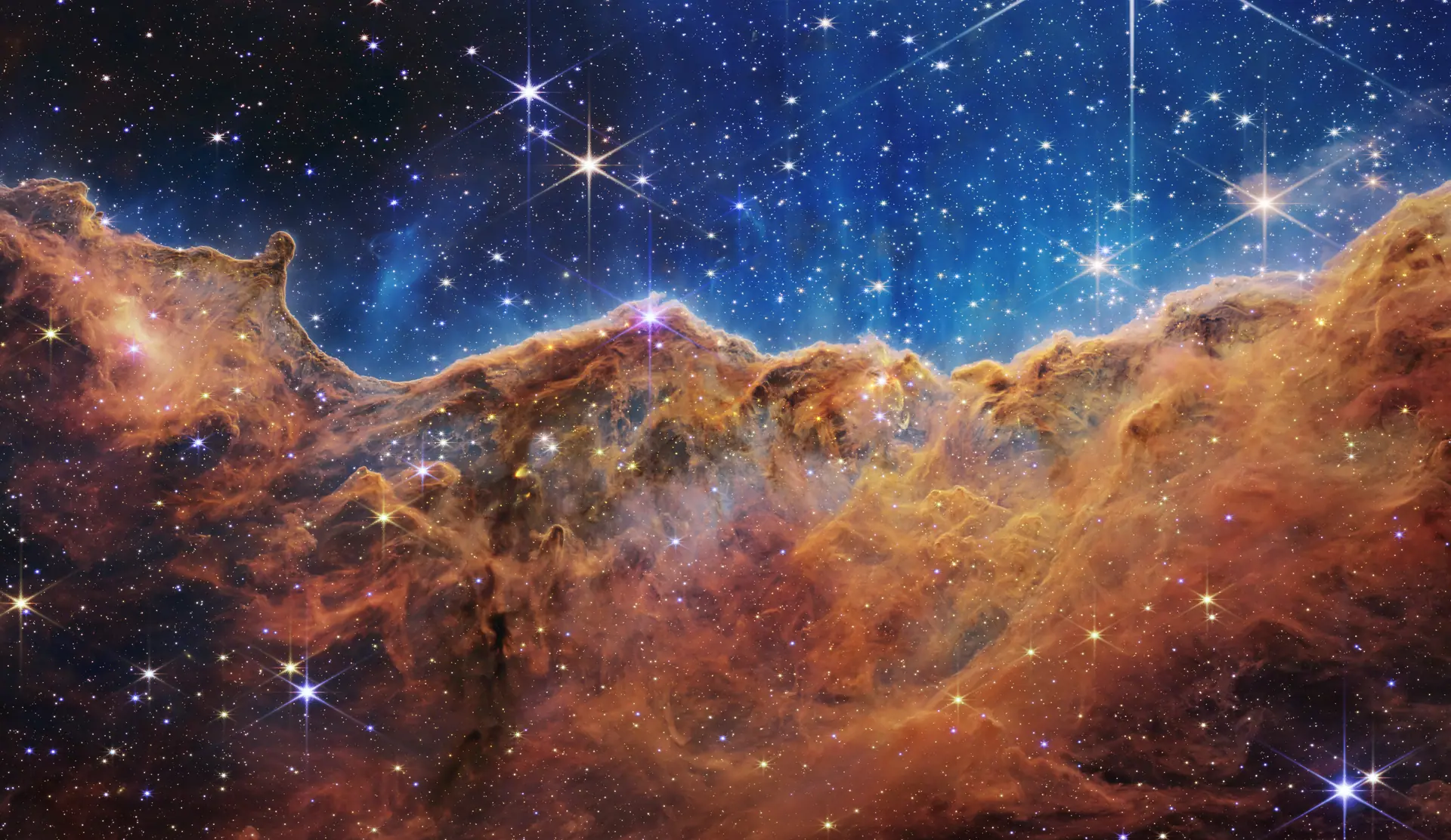 The Cosmic Cliffs, a glowing amber wall of gas along the edge of a star-forming cavity in the Carina Nebula